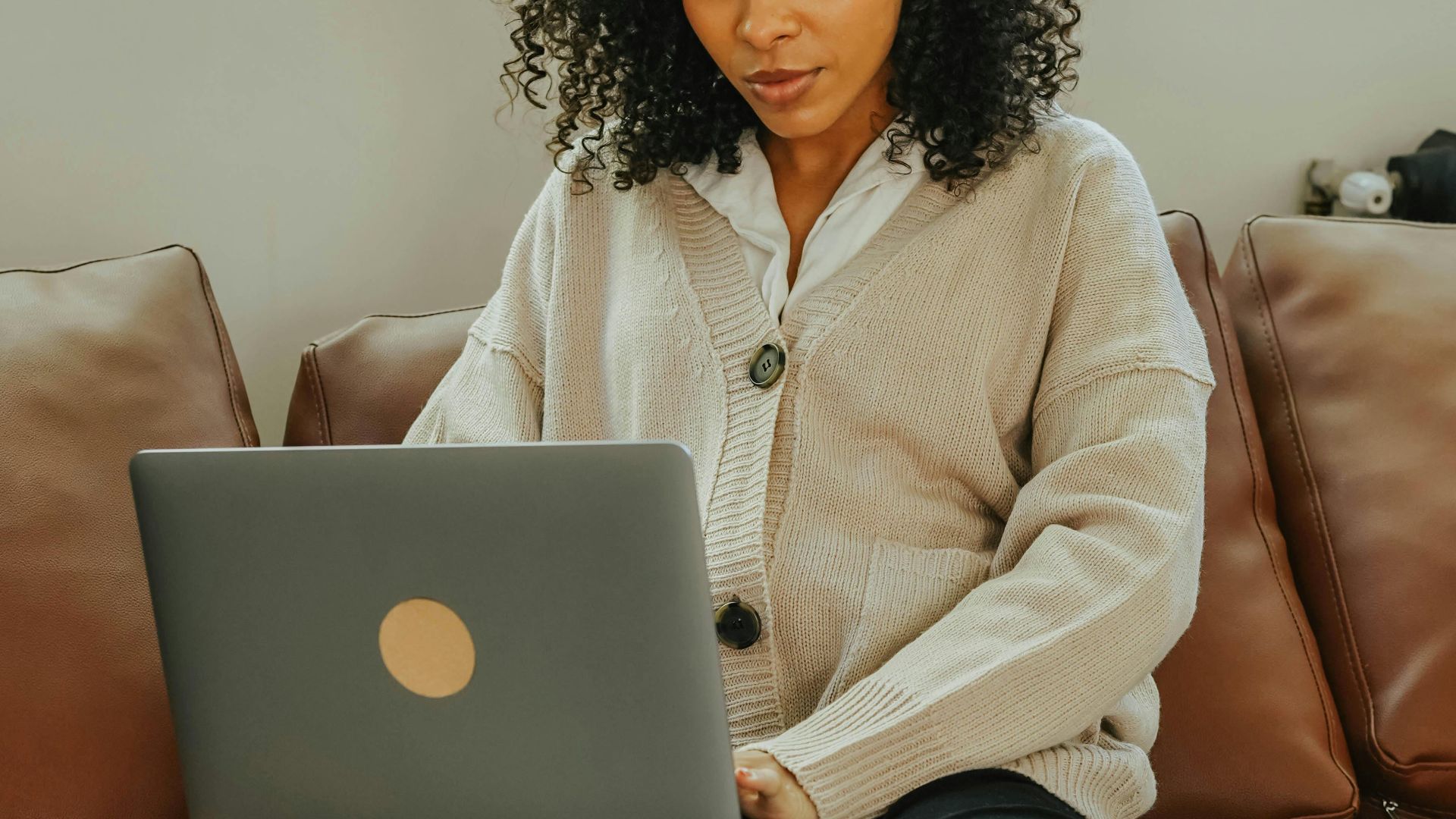 Focused woman with curly hair using a laptop on a brown leather couch indoors.
