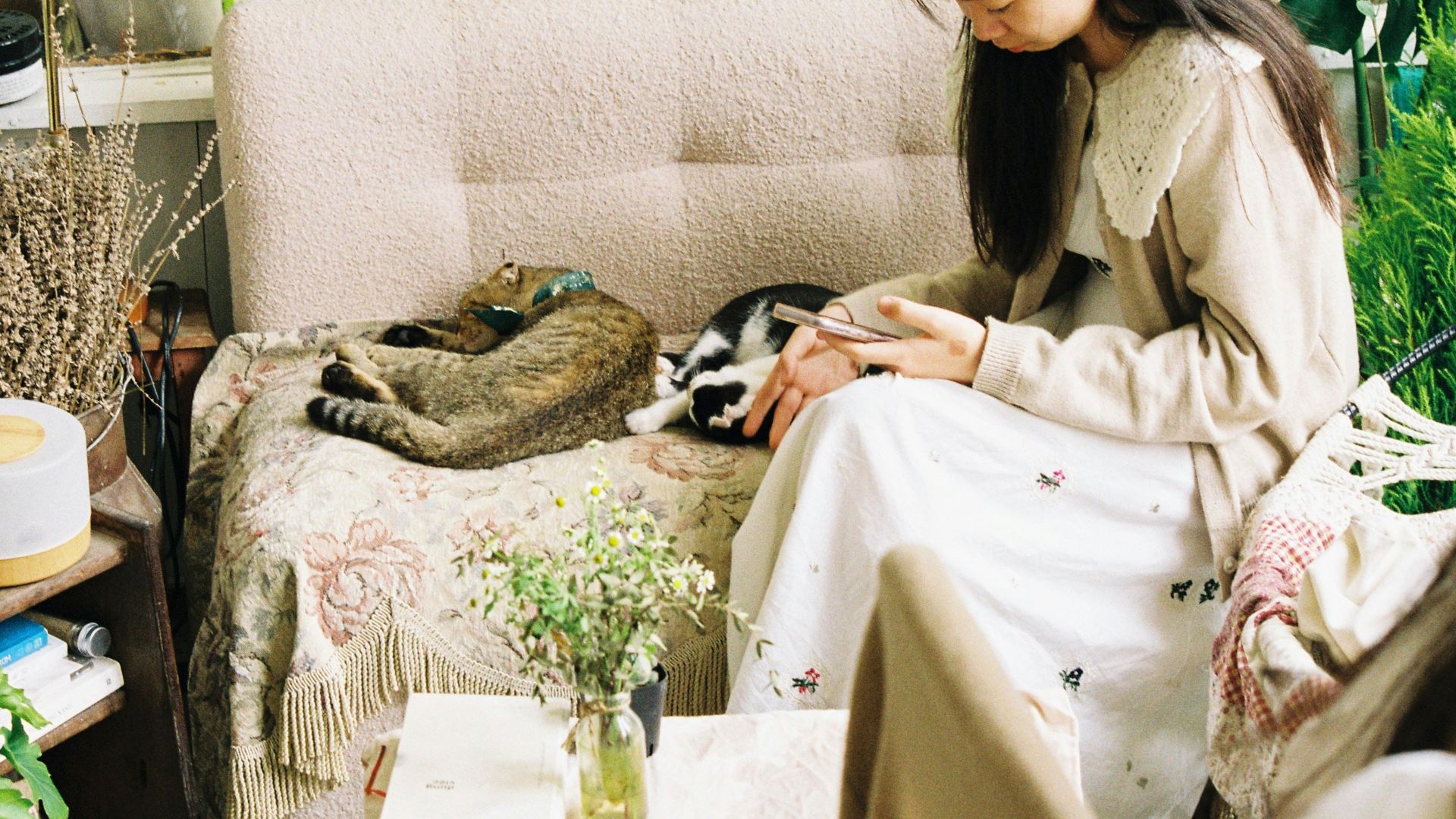 A young woman enjoys a peaceful moment with her cats on a sofa in a lush indoor garden setting.