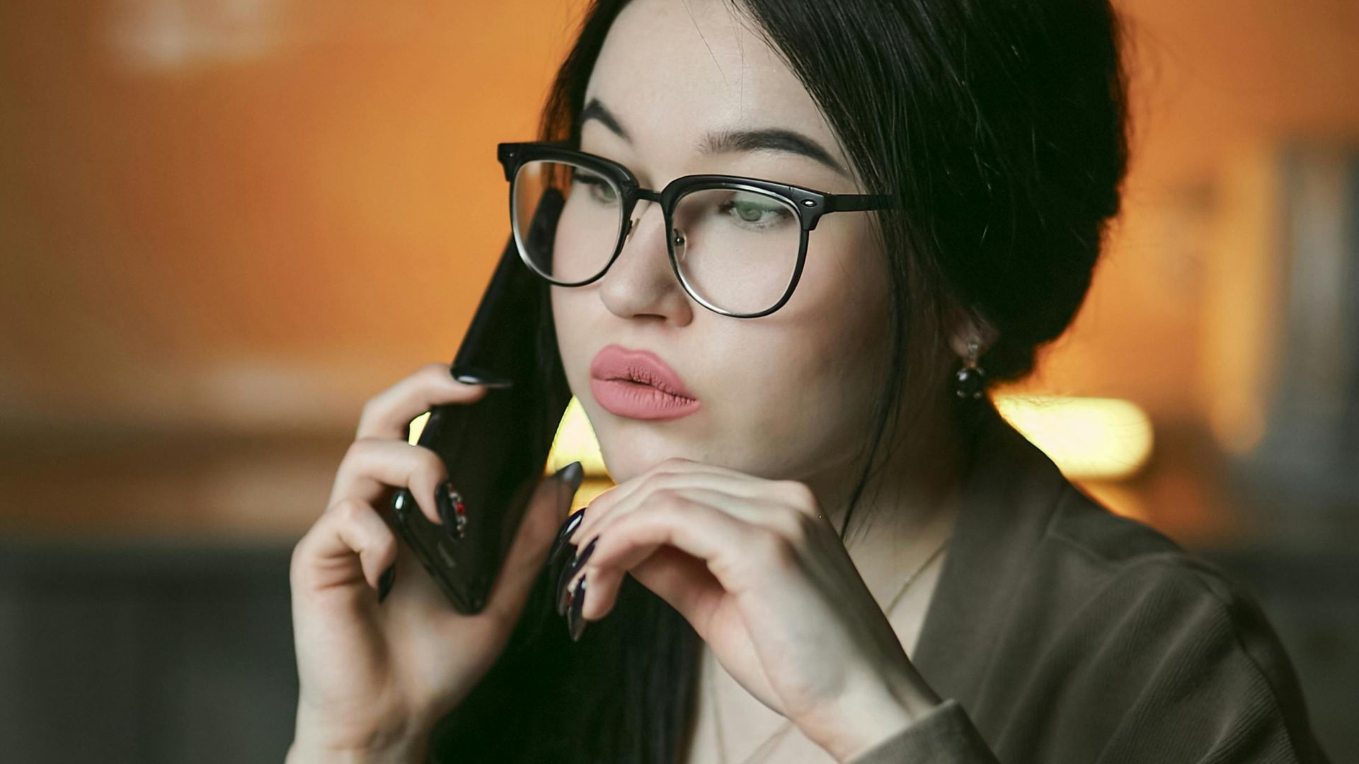 A woman with eyeglasses engaged in a phone call indoors, focused and thoughtful.
