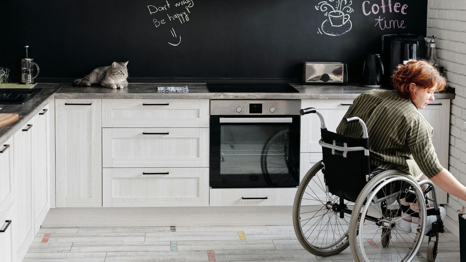 Woman in wheelchair using modern kitchen with cat and chalkboard wall. Warm and inviting atmosphere.