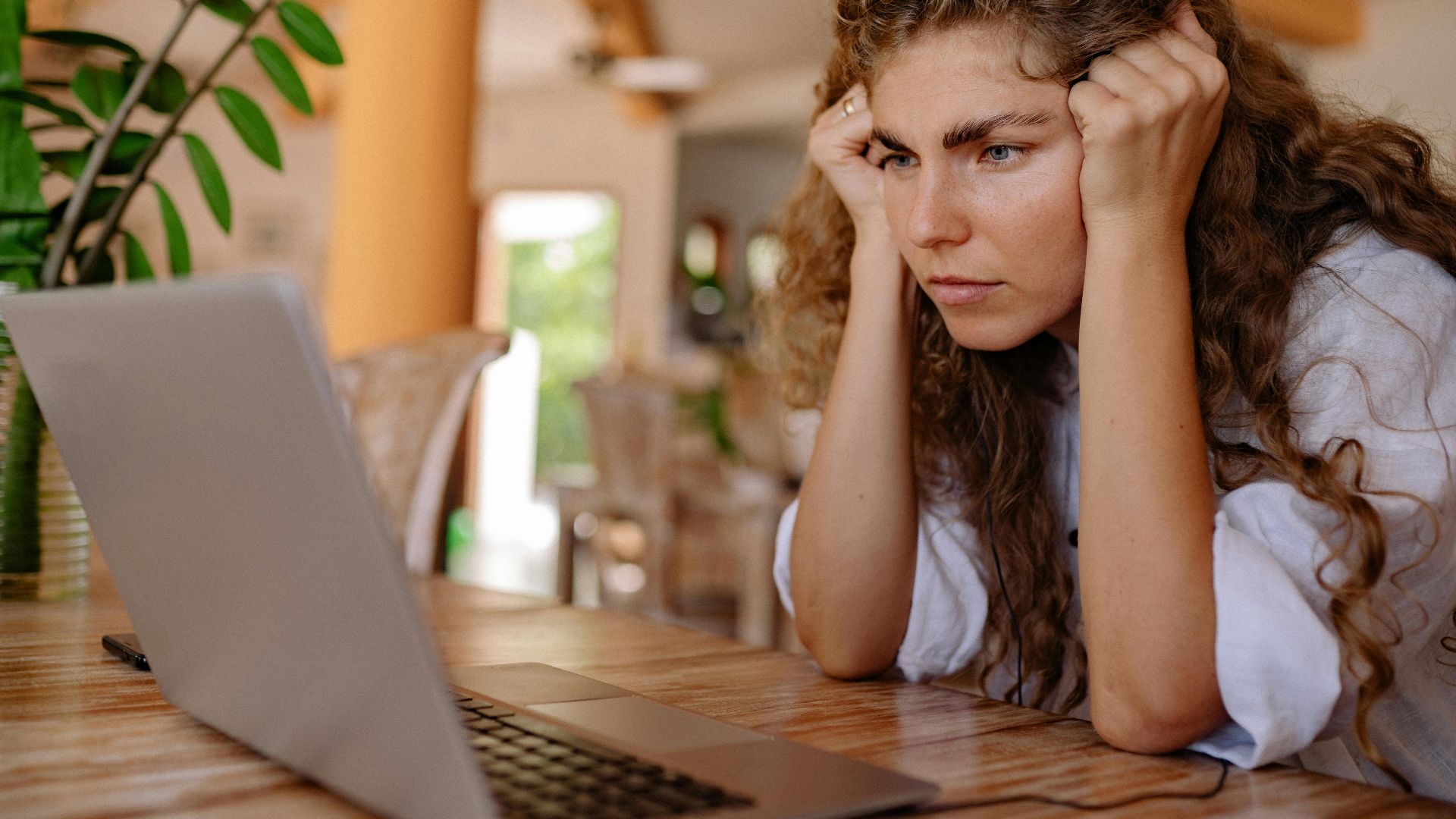 A woman with curly hair intensely focusing on her laptop indoors with plants and natural lighting.