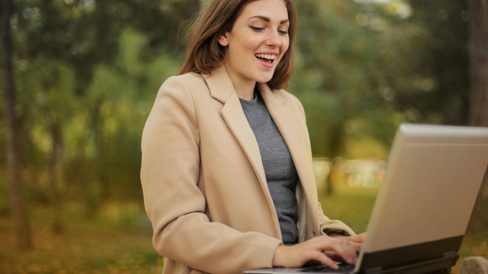 Smiling woman using laptop for remote work in a scenic autumn park setting.