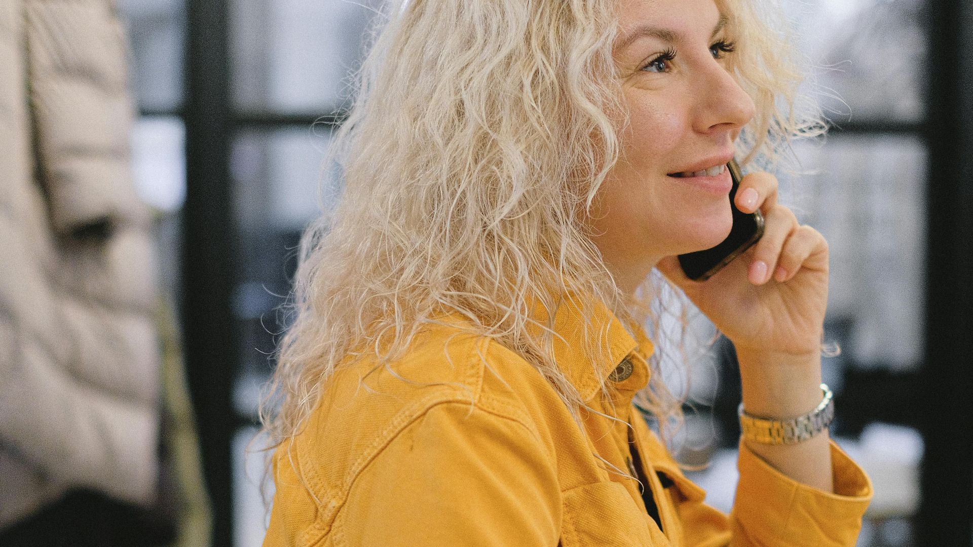 Side view of positive businesswoman sitting at table and writing notes while having conversation on mobile phone