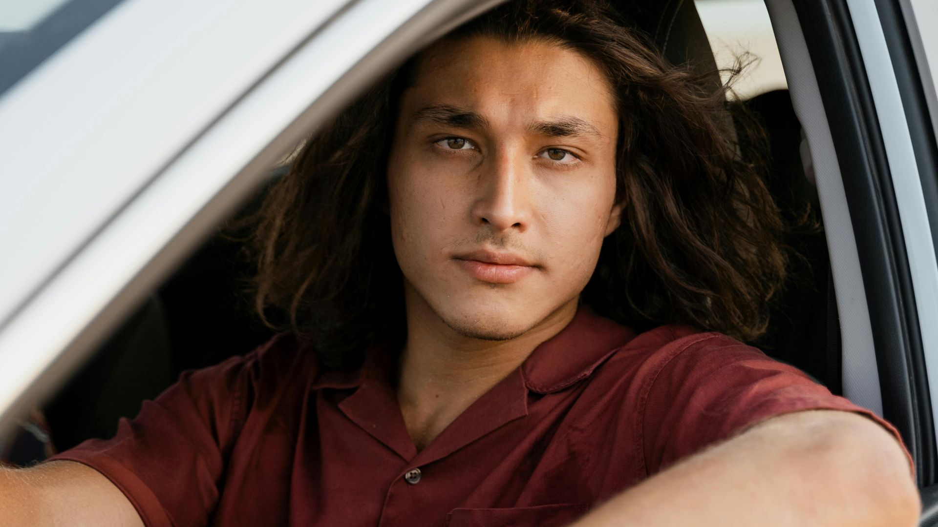 Portrait of a young man in a red shirt, sitting inside a car, ready to drive.