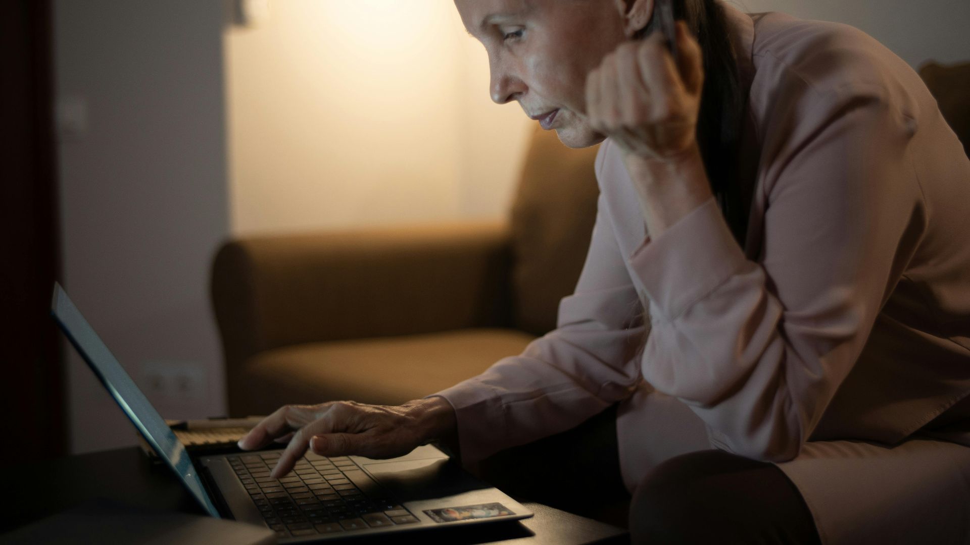 Senior woman using laptop in a dimly lit living room, focused on screen.