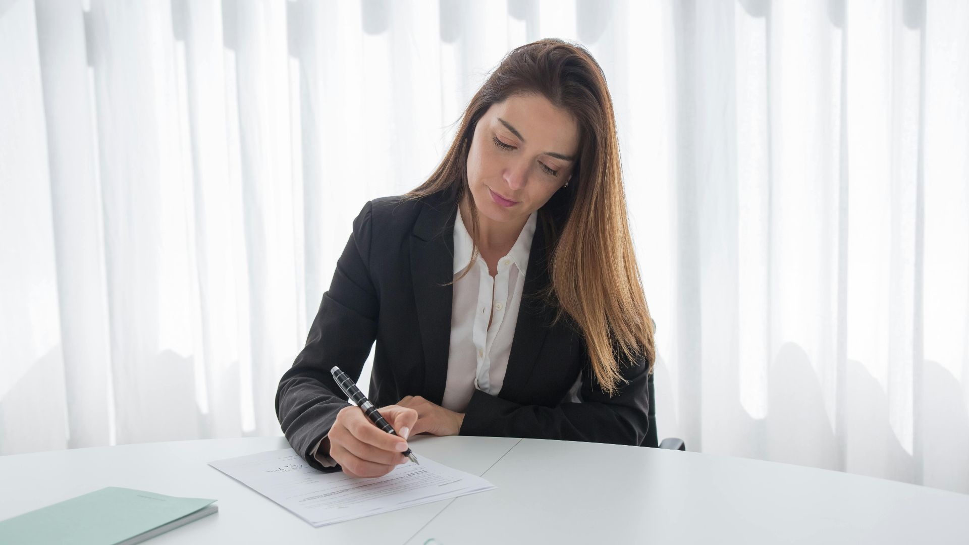 A professional woman in a suit signing paperwork at a desk, showcasing corporate elegance.