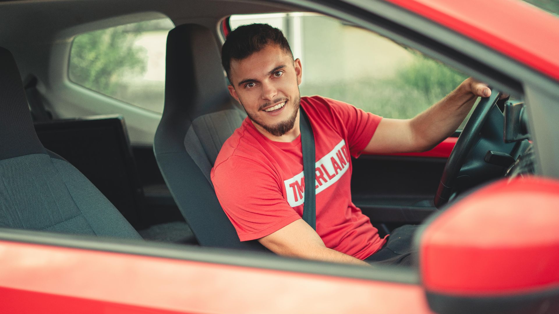Smiling man driving a red car while wearing a seatbelt, showcasing safety and joy.