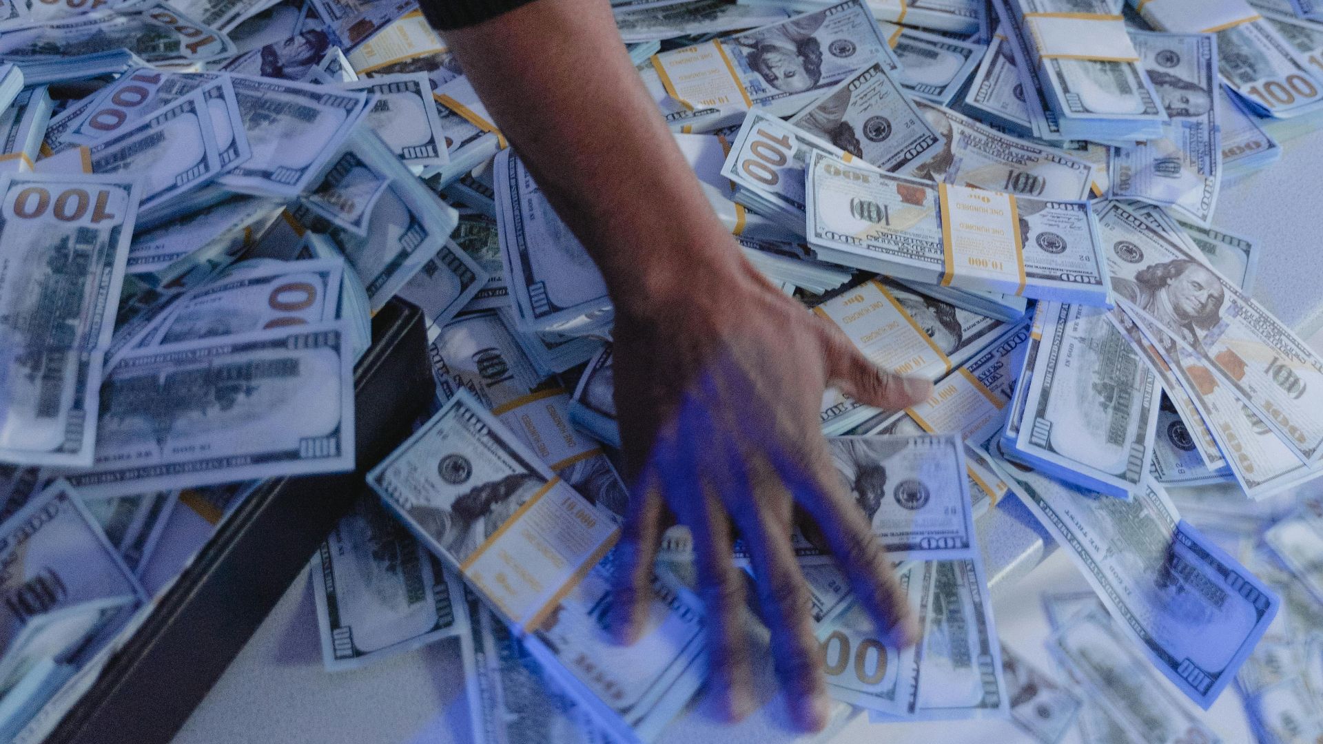 Adult man organizing a massive pile of US dollar bills indoors in dynamic lighting.