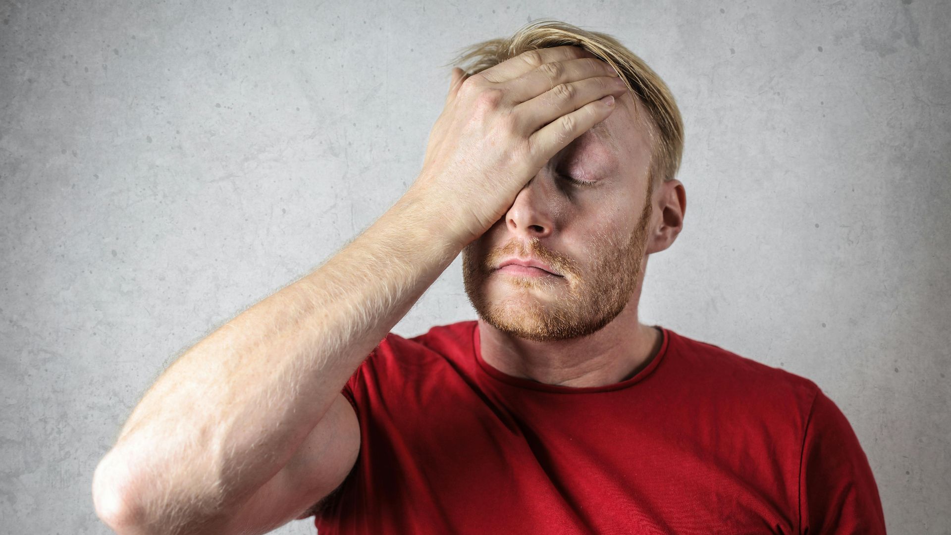 A frustrated man in a red shirt holds his head in stress against a neutral background.