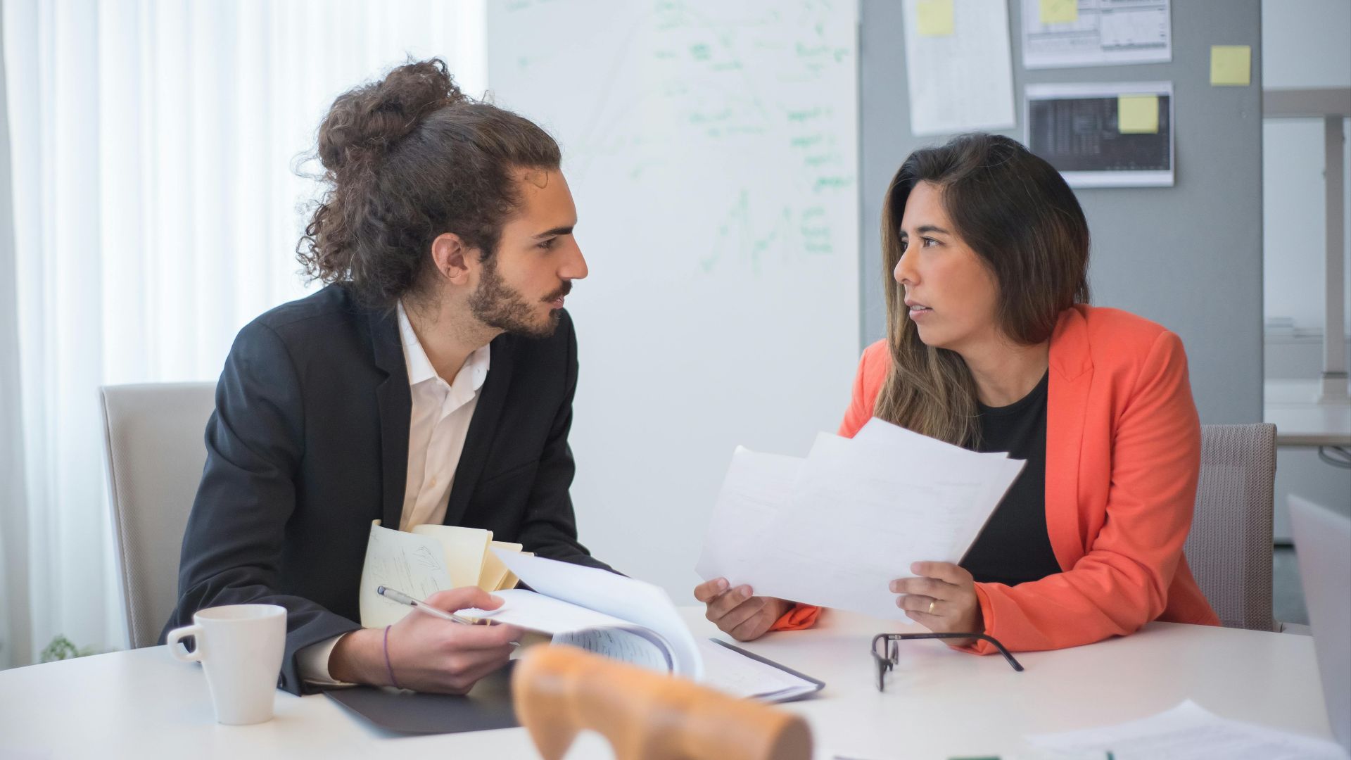 Two colleagues engaged in a discussion at an office table with documents and coffee.
