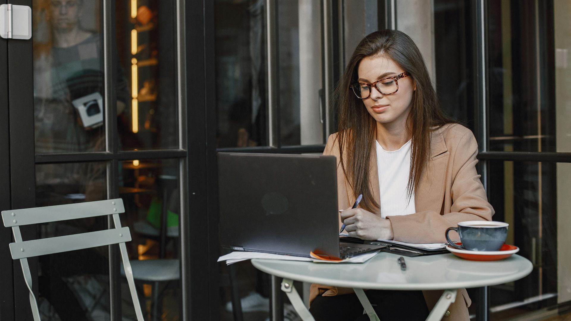 Woman in business attire working remotely on laptop at cafe table outdoors.
