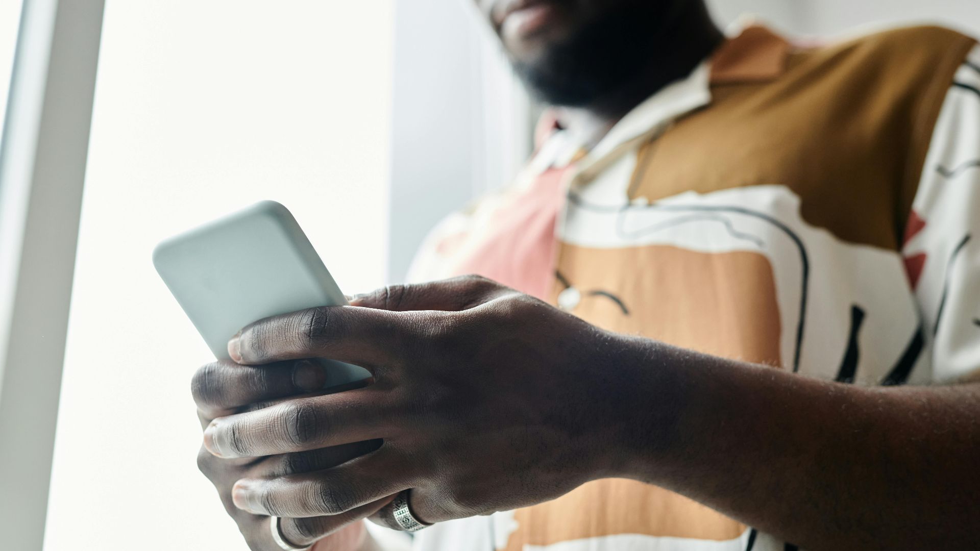 A close-up of a man using a smartphone by a window, featuring a modern patterned shirt.