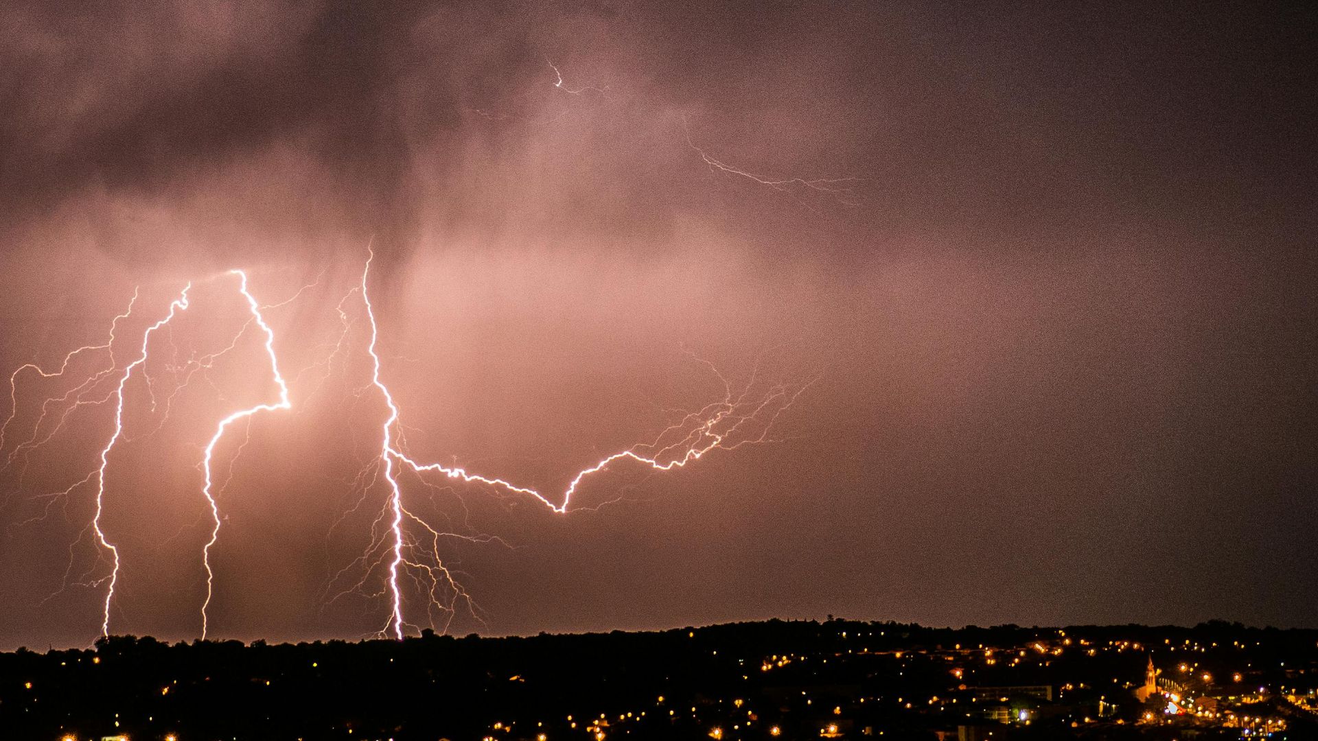 A powerful lightning storm illuminates the night sky over a cityscape, creating a dramatic scene.