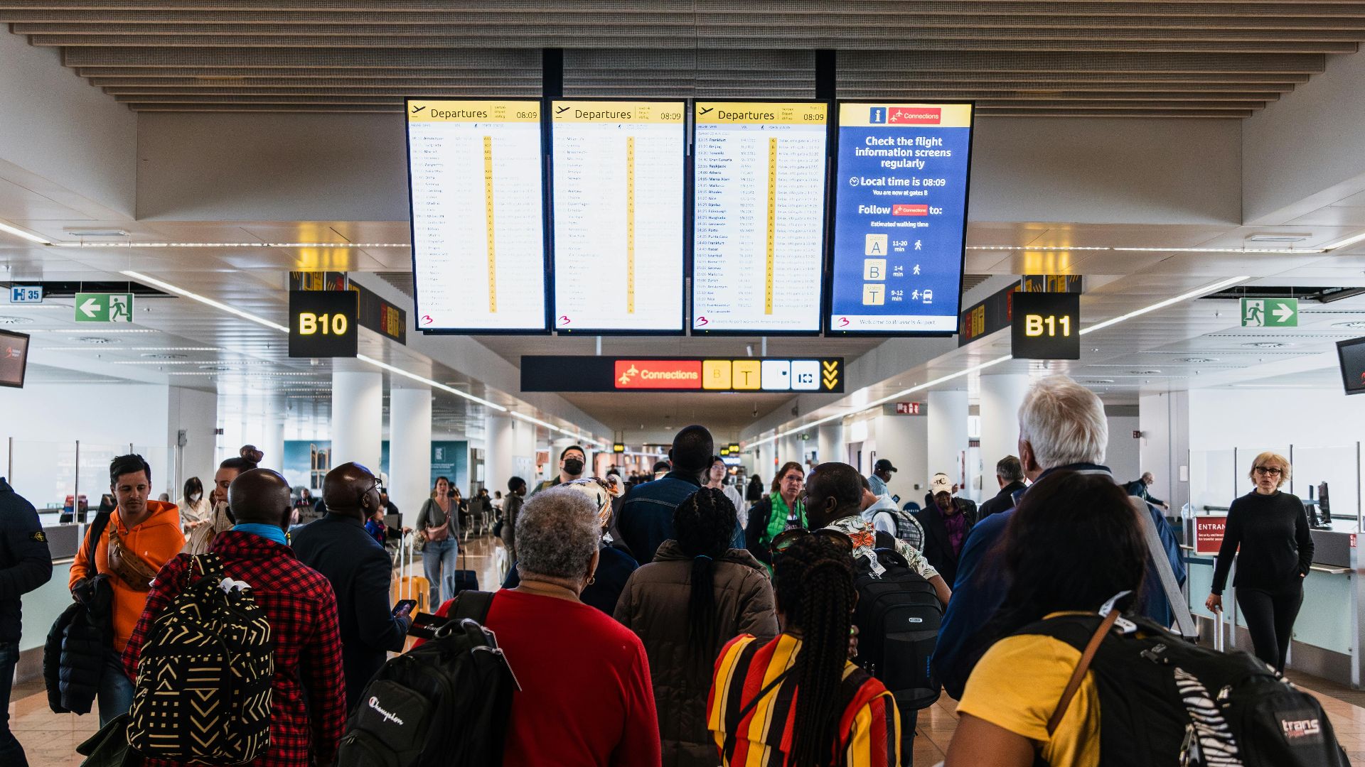 Travelers lining up in a busy airport terminal with baggage under flight information screens.