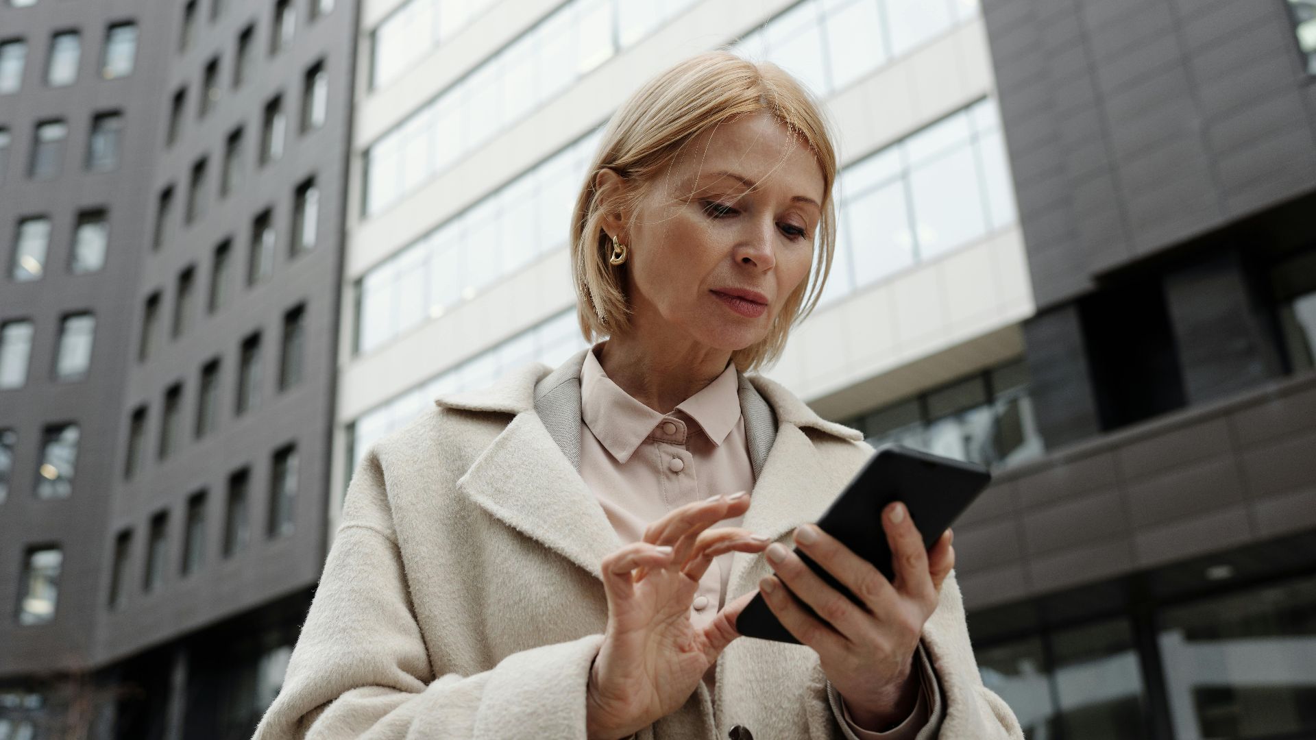 Woman in coat using smartphone in front of modern building. Professional and focused expression.