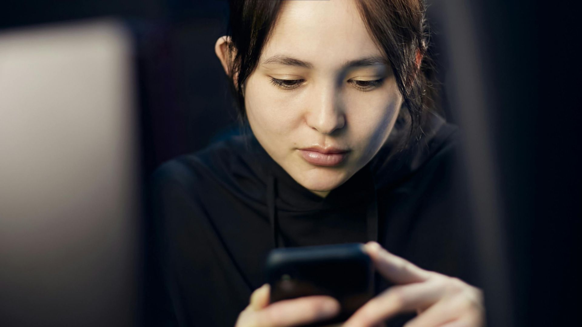 A woman intently looking at her smartphone while seated indoors, dim lighting.