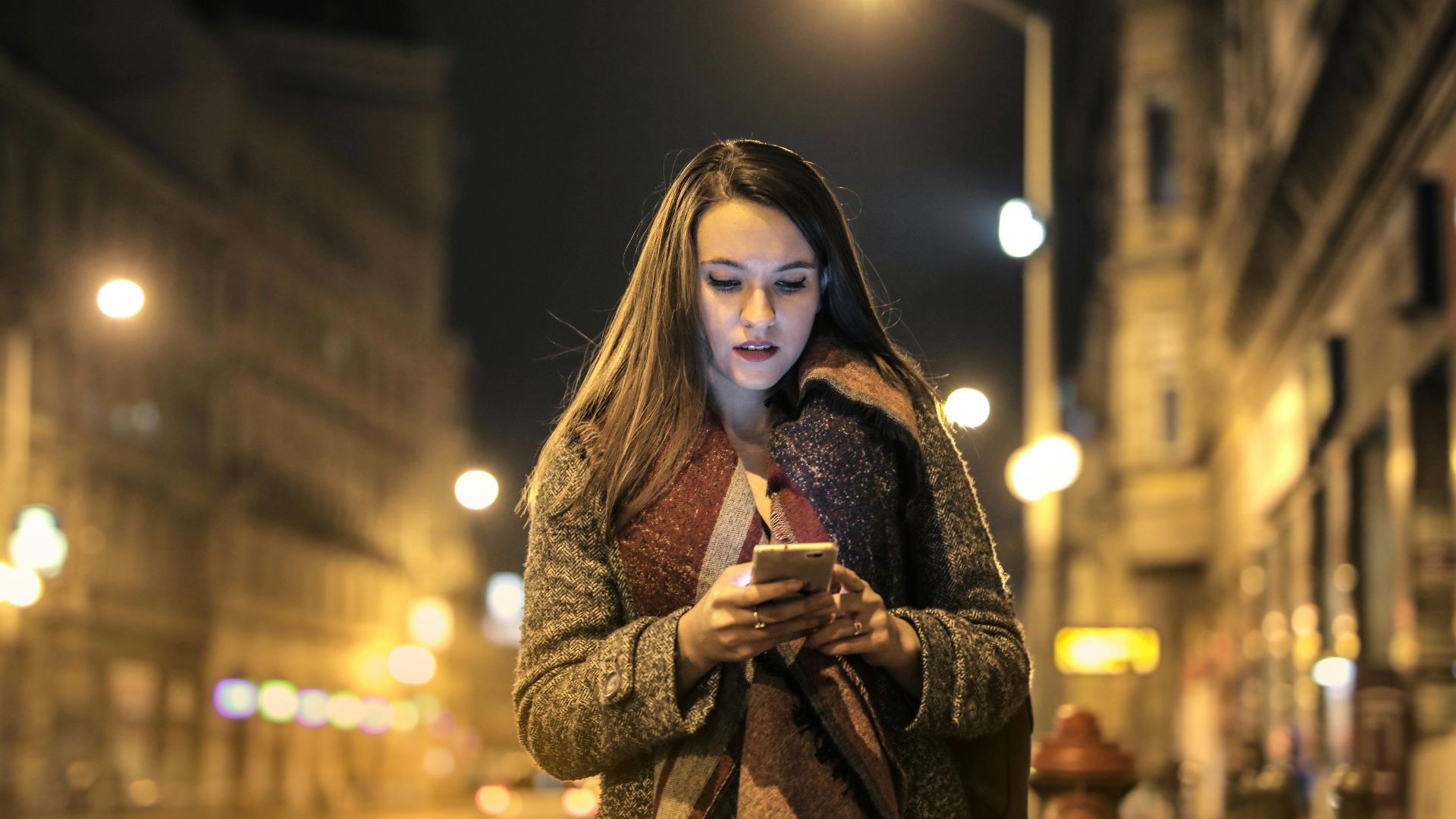 Elegant woman walks through urban street at night, engaged with her smartphone, surrounded by city lights.