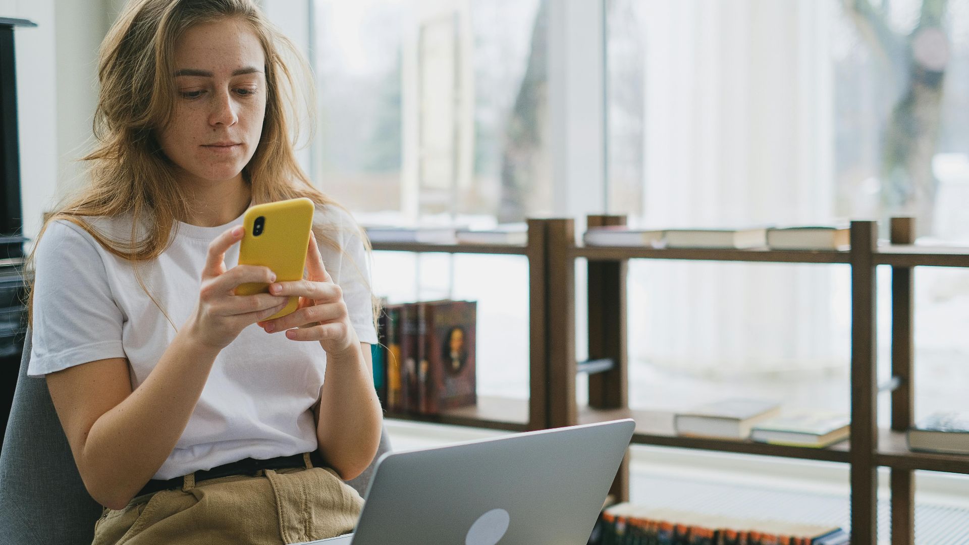 Young woman multitasking with smartphone and laptop in a cozy indoor setting.