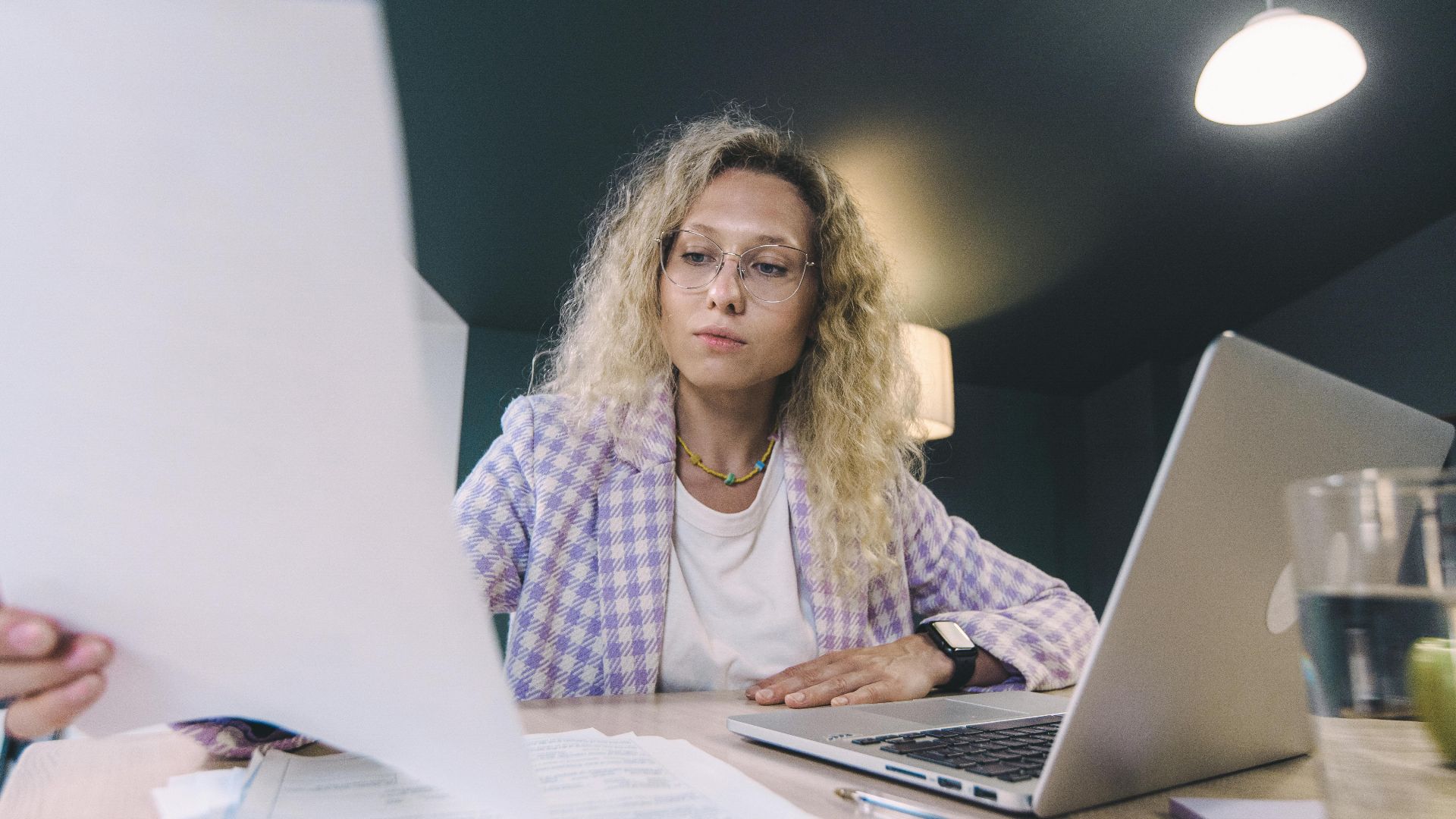 Woman in an office environment reviewing documents with focus, surrounded by technology.