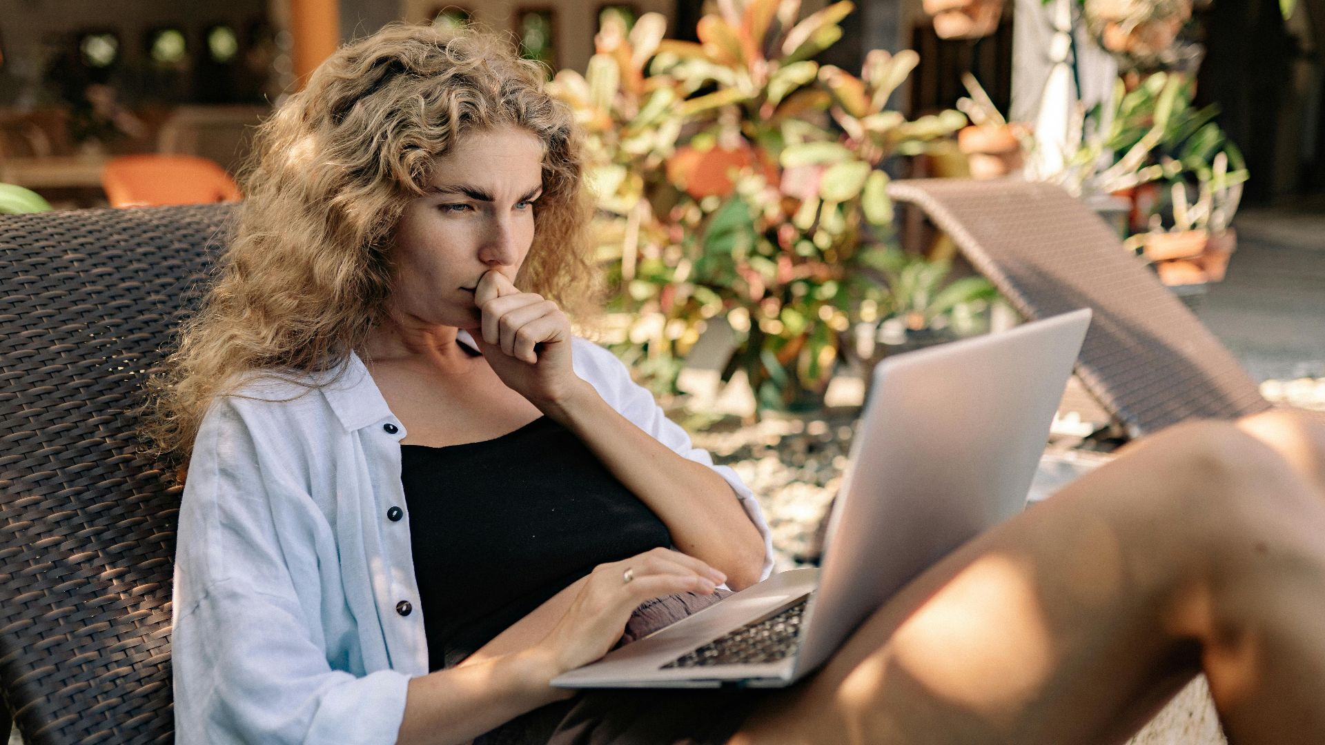 Woman with curly hair working remotely on a laptop while lounging outdoors in a tropical setting.