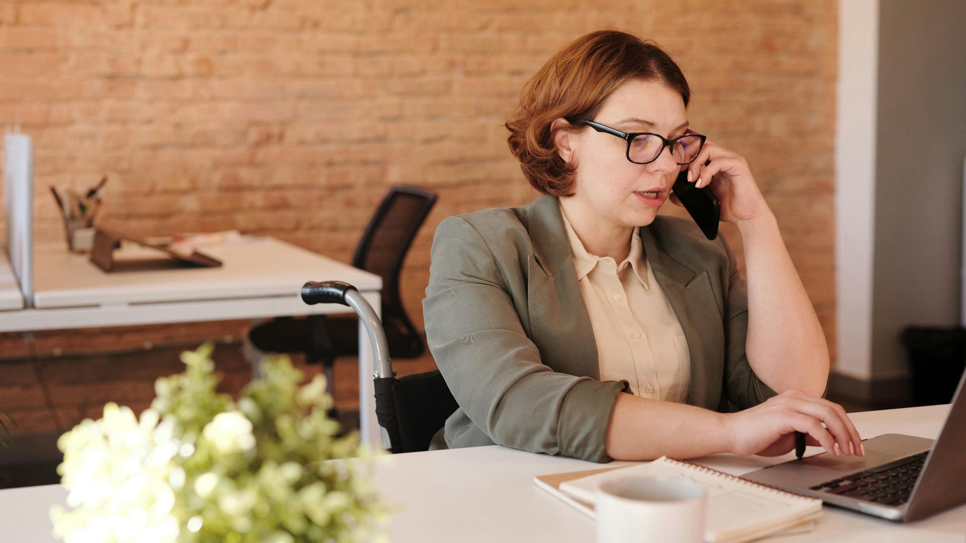 Professional woman in wheelchair working remotely on laptop and phone in a modern office.