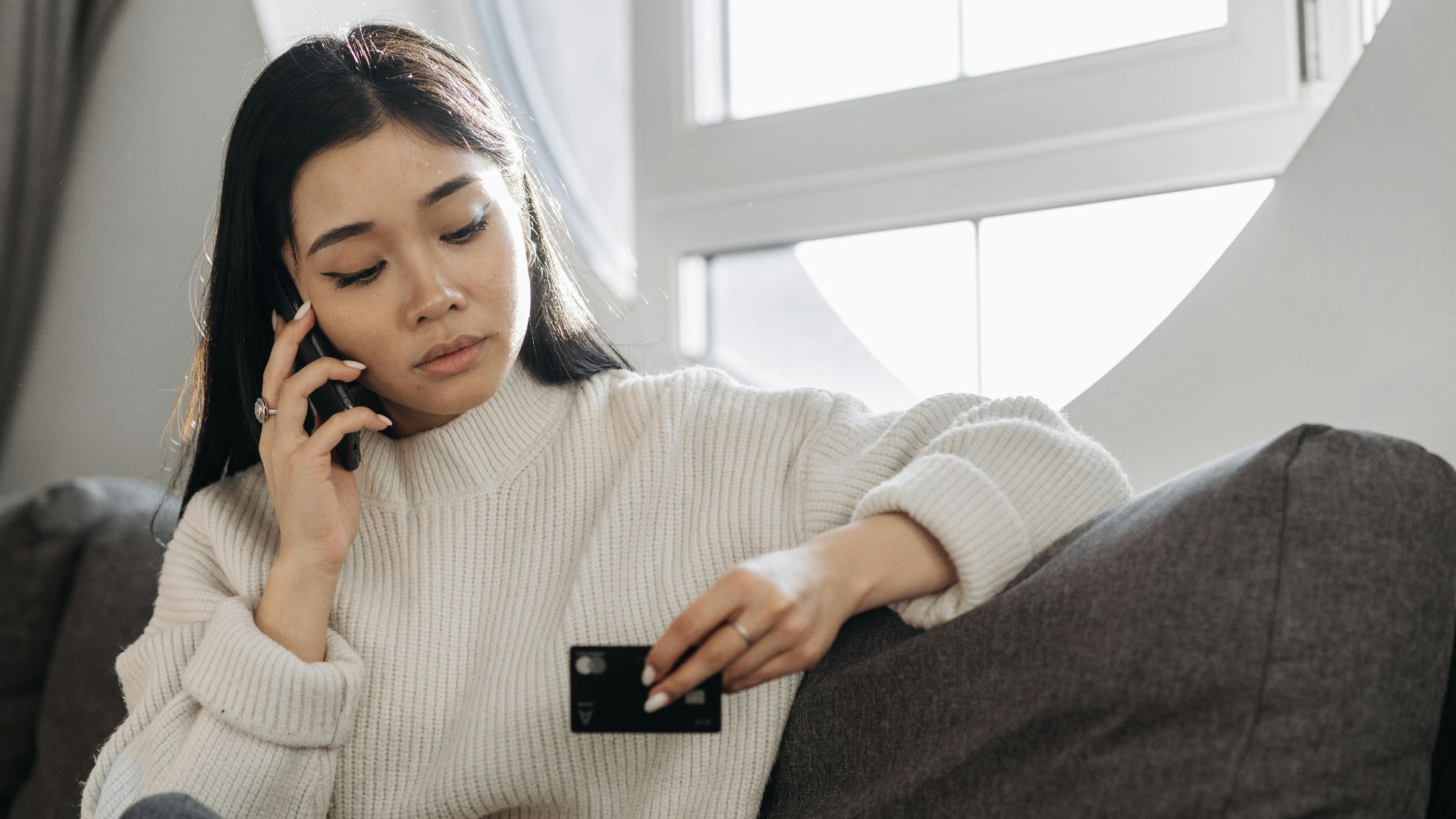 A woman sits on a sofa, multitasking with a phone and credit card, focusing on online shopping.