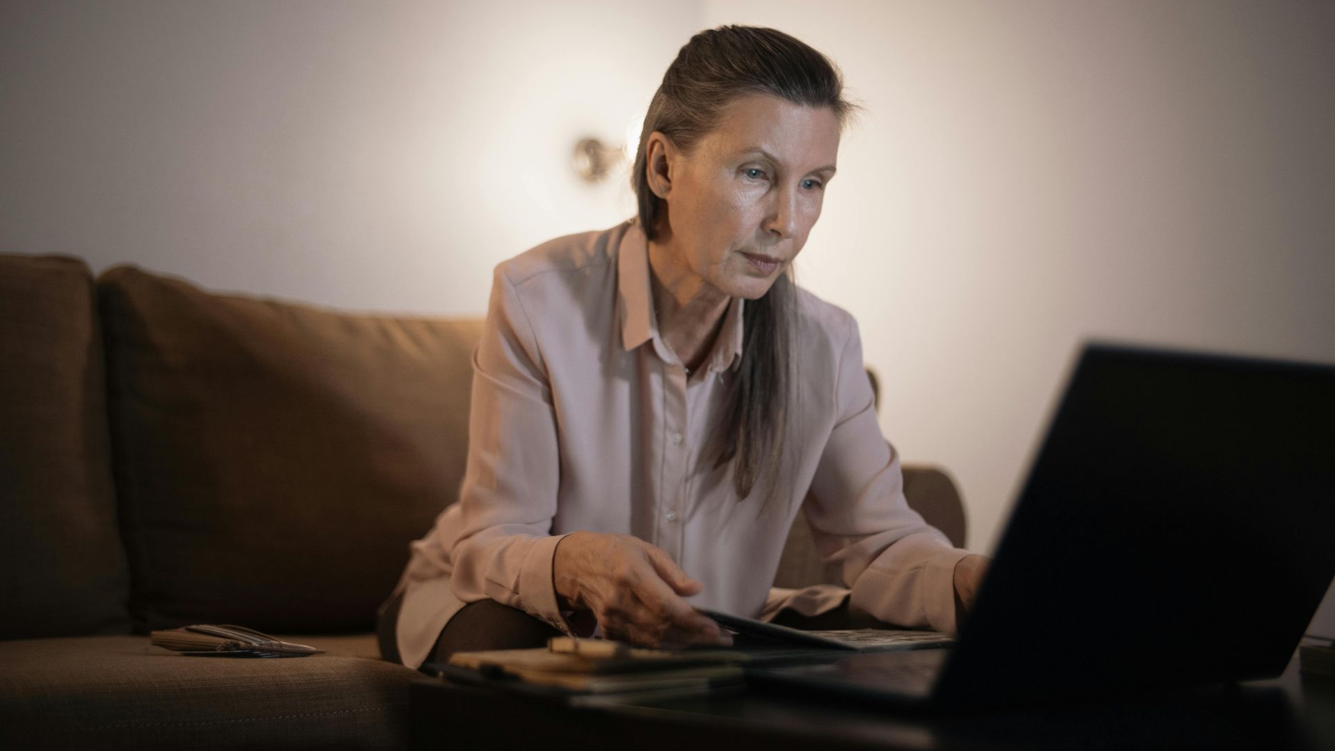 Elderly woman working on laptop in dimly lit living room setting, focused and engaged.