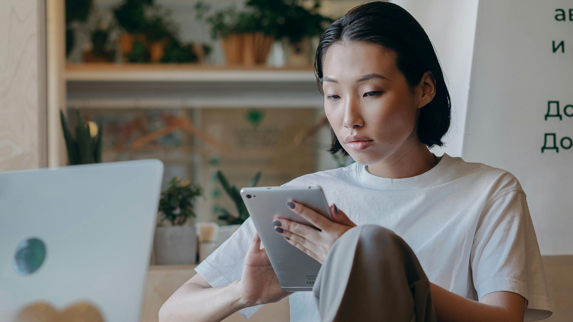 Focused Asian businesswoman using a tablet in a modern, plant-filled workspace with natural lighting.