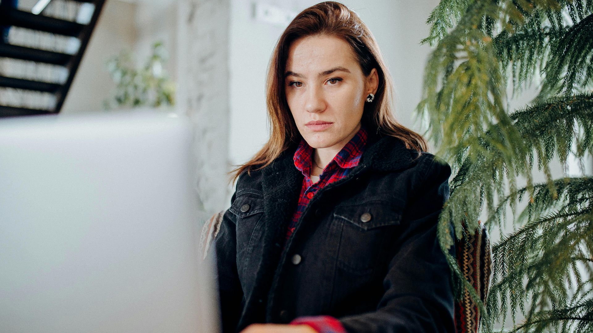 Young woman wearing a denim jacket working intently on a laptop in a modern indoor setting.