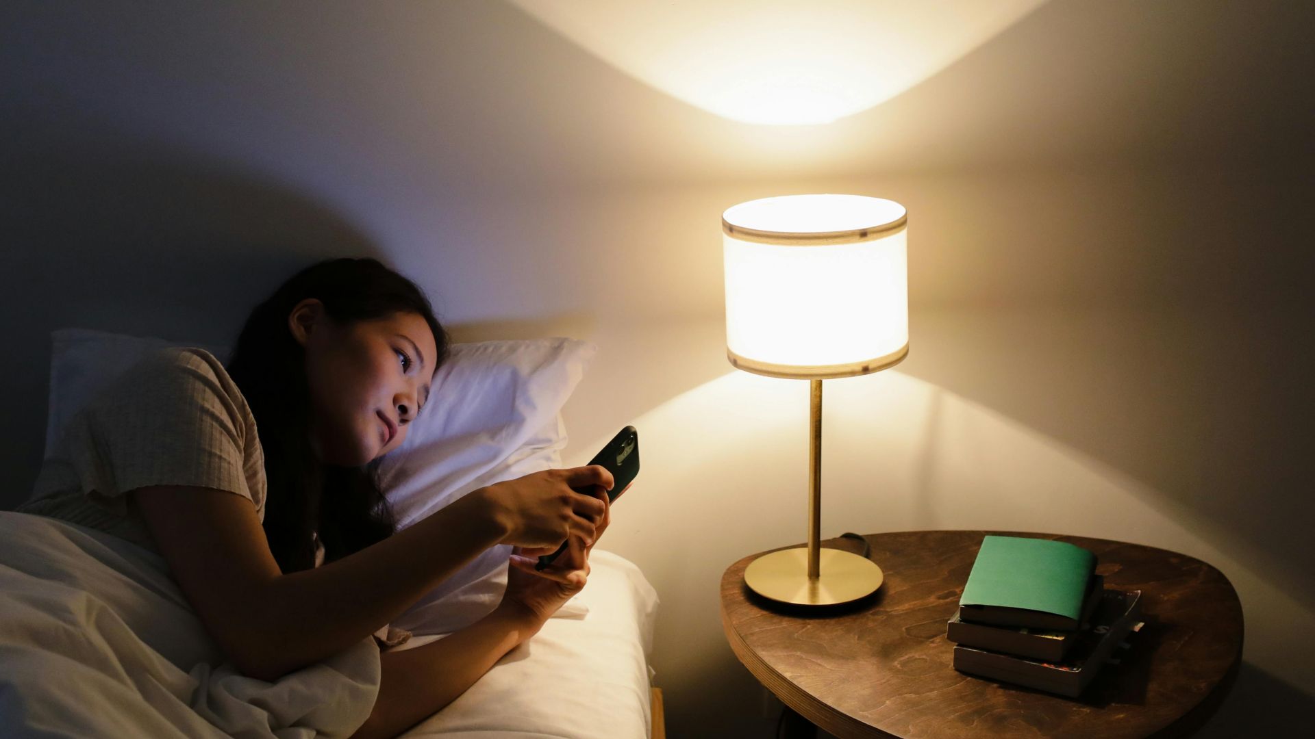 A woman relaxes in bed, using her smartphone beside a lit table lamp in a cozy room.