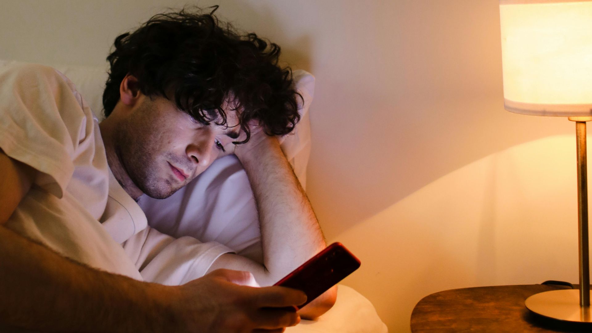 A man lying in bed using a smartphone beside a glowing bedside lamp.