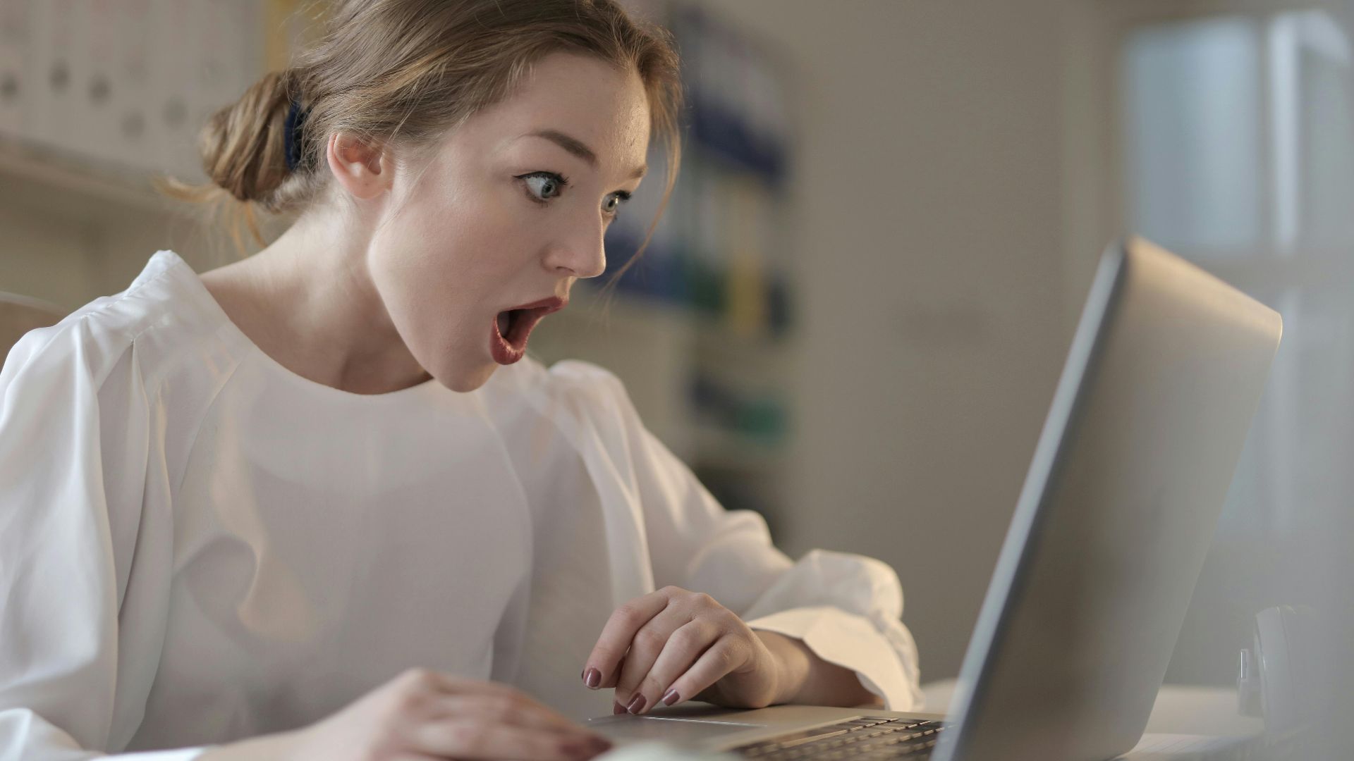 Surprised woman sitting at desk with laptop indoors, expressing amazement.
