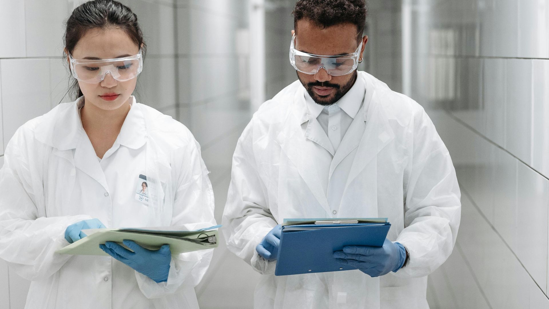 Two researchers in lab coats review documents in a clinical laboratory hallway.