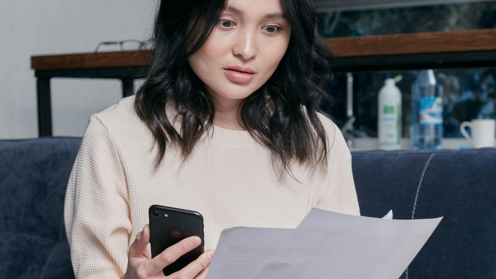 Young woman reviews documents and phone indoors, showcasing concern while seated on a couch with scattered papers. Business concept.