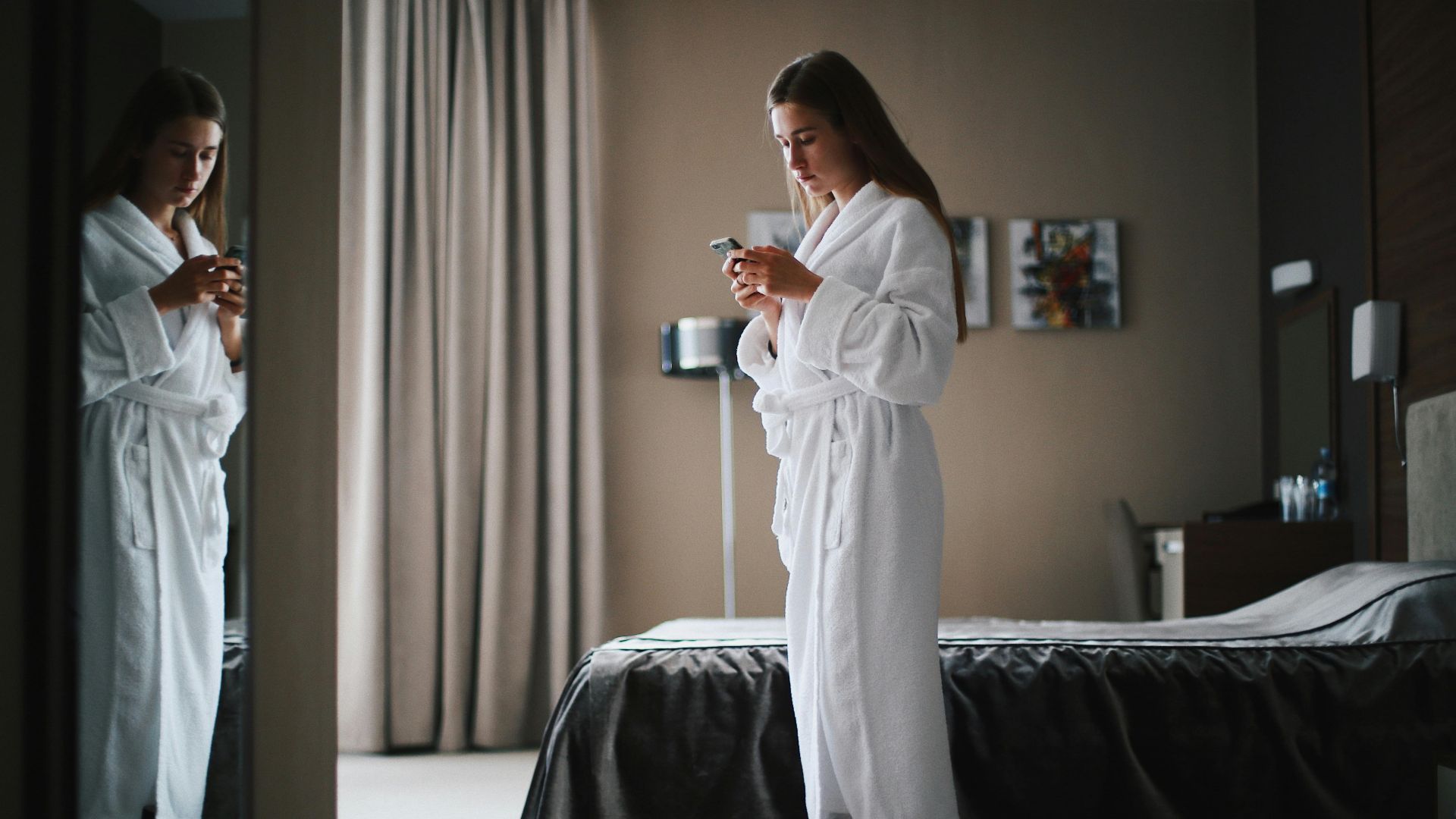 Woman in bathrobe looking at smartphone in a modern hotel room, near a mirror.