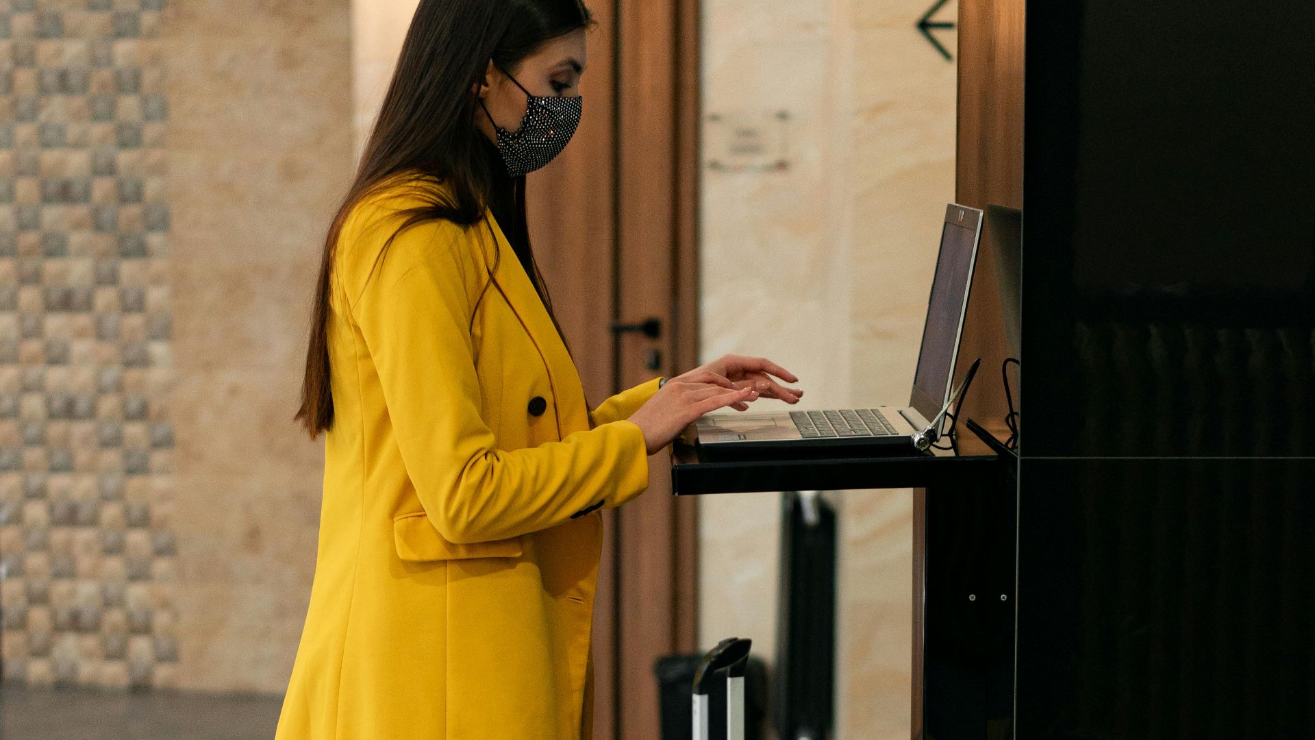 Business traveler using laptop at hotel reception with yellow suitcase.