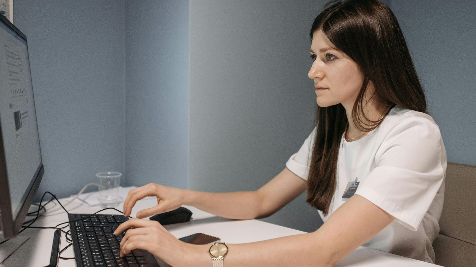 Focused woman in white uniform typing on computer in office setting.