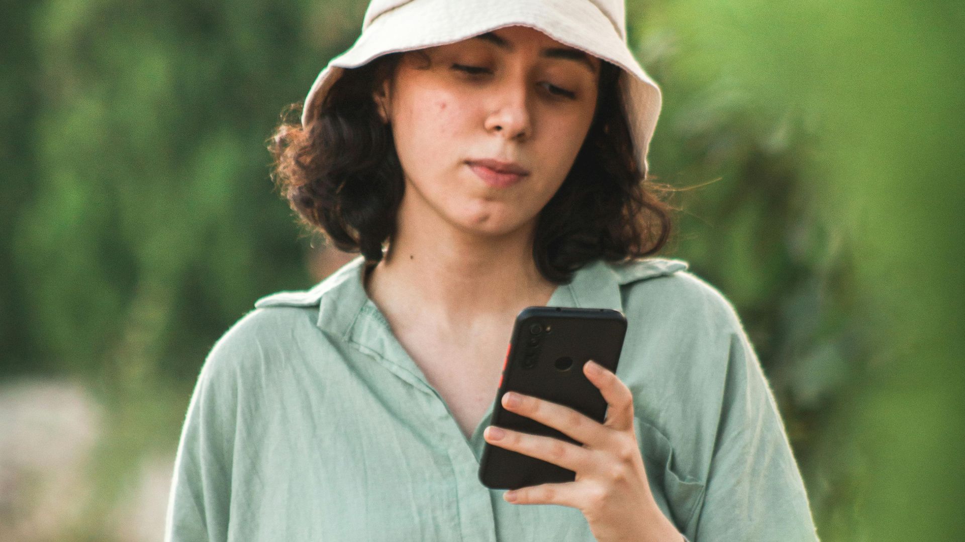 A young woman in a bucket hat uses her smartphone while walking outdoors.
