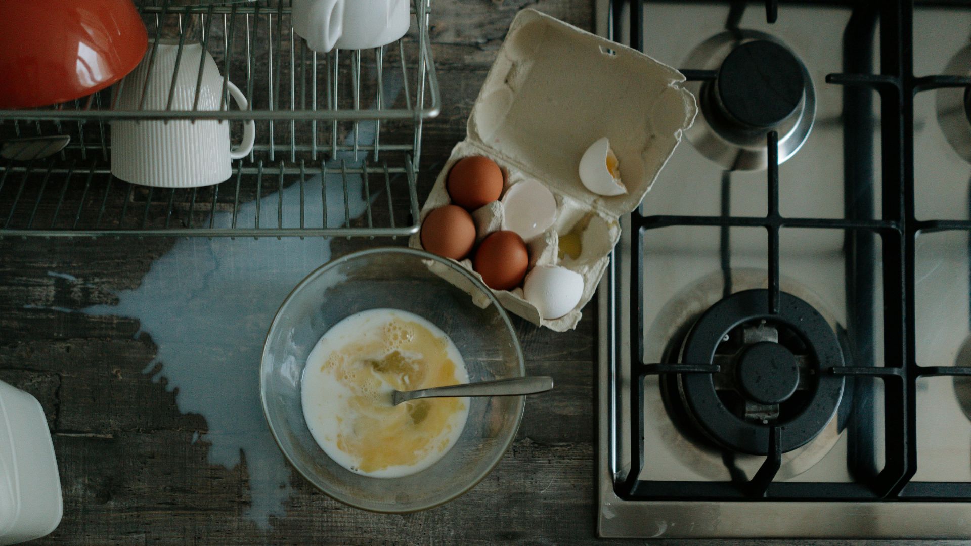 Eggs and milk spilled on a kitchen counter next to a stove and dishrack.