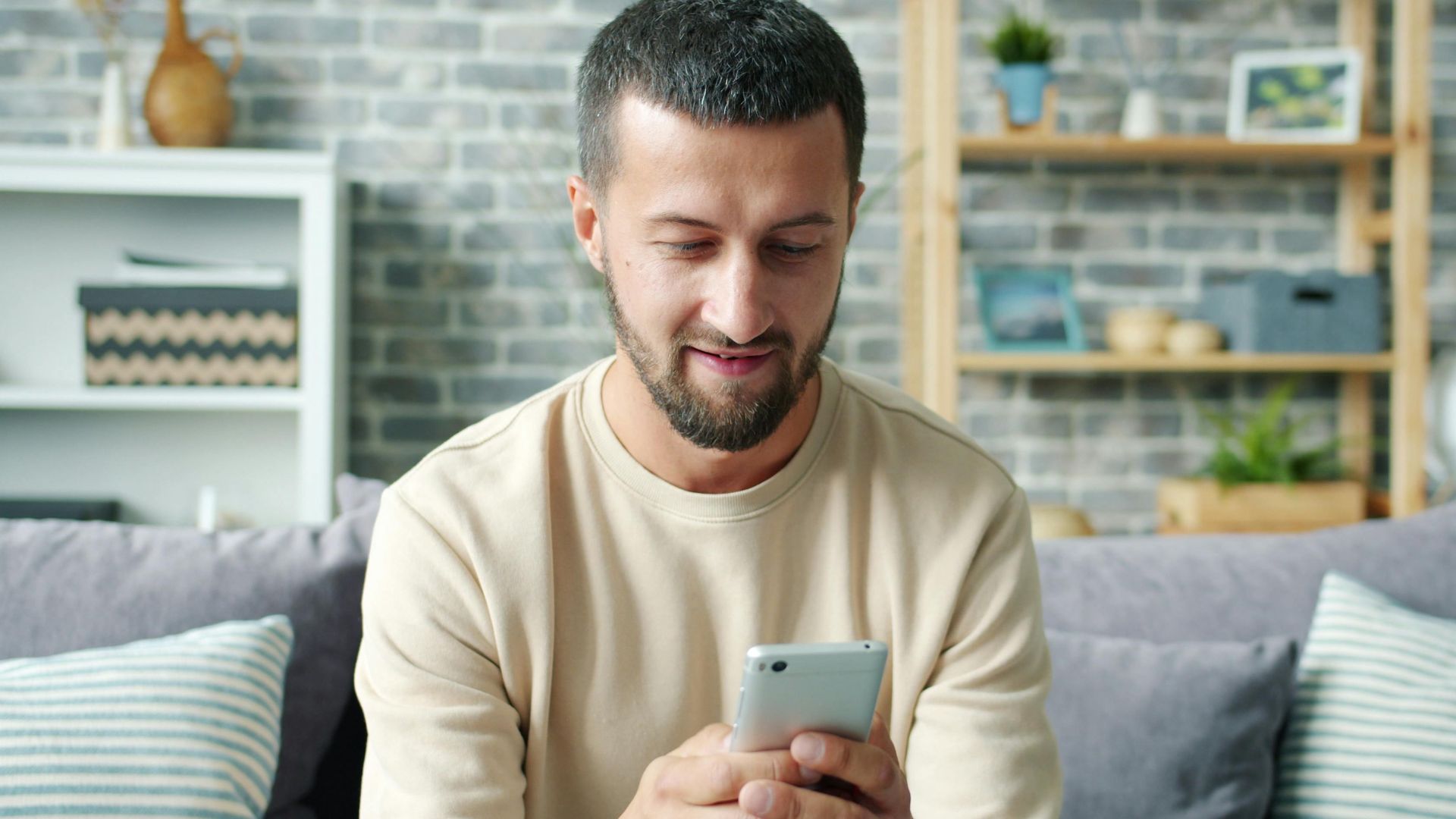 Casually dressed man using smartphone while sitting on a cozy sofa in a modern living room.
