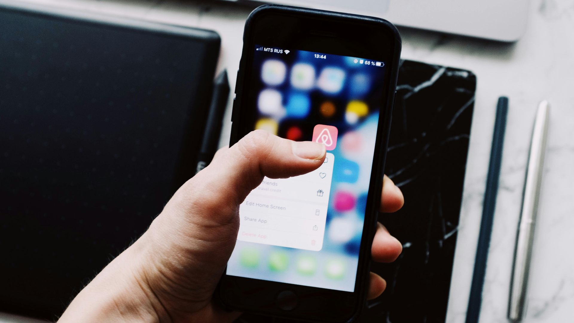 A person holds a smartphone displaying app icons over a desk with digital tablets and pens.