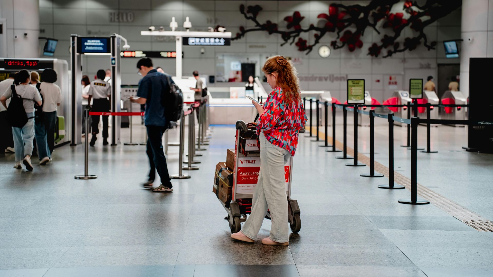 A traveler checks her phone while waiting with a luggage cart at a busy airport terminal in Guangzhou.