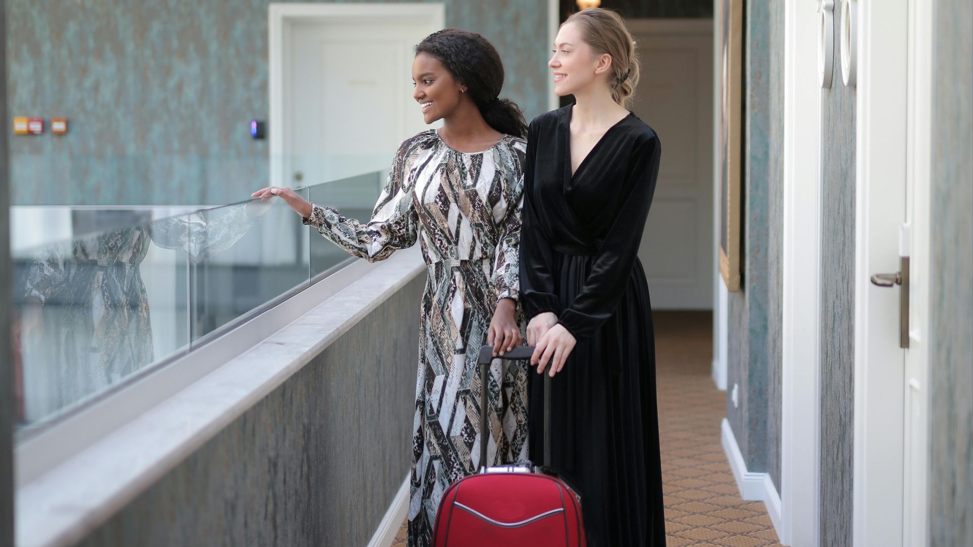 Two fashionable women with luggage in a stylish hotel hallway.