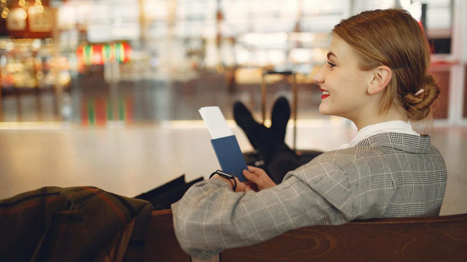 Side view of cheerful female student in checkered jacket smiling away while chilling in hall with outstretched legs and passport in hands