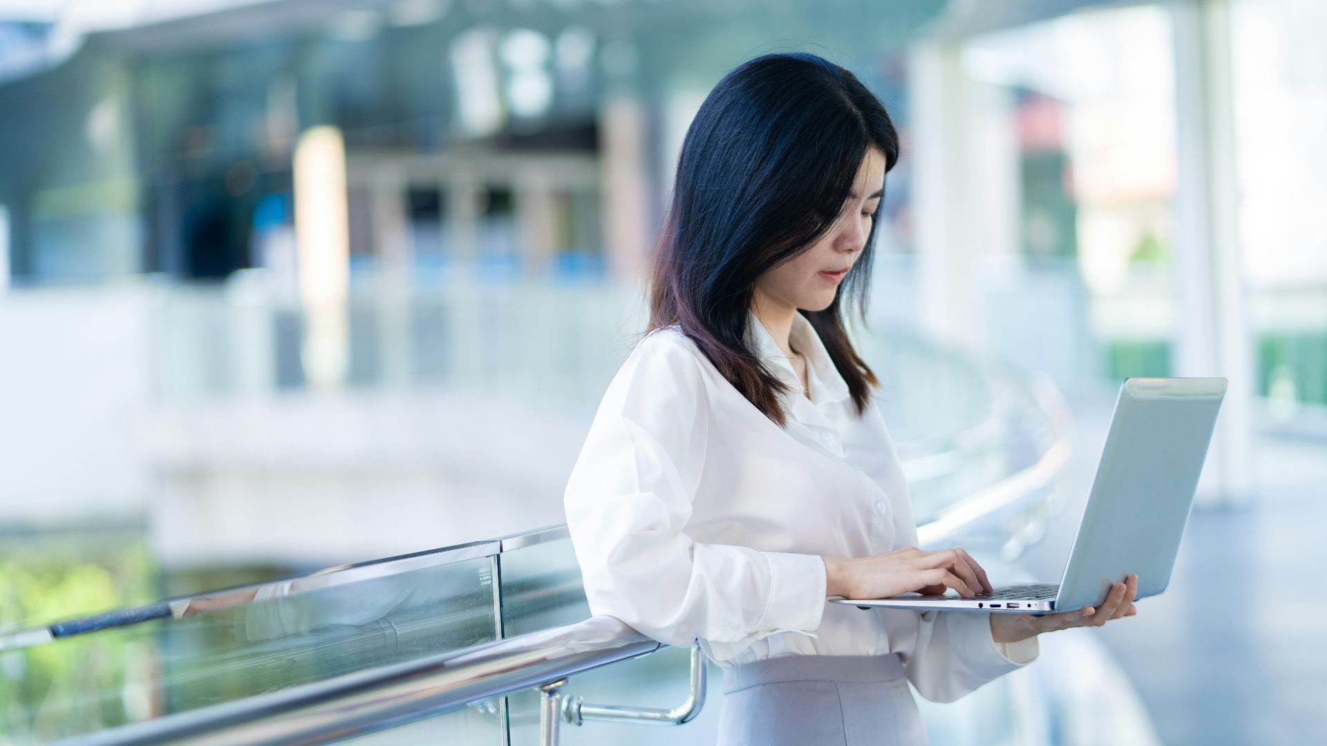 Asian businesswoman multitasks on a laptop in a modern office setting.