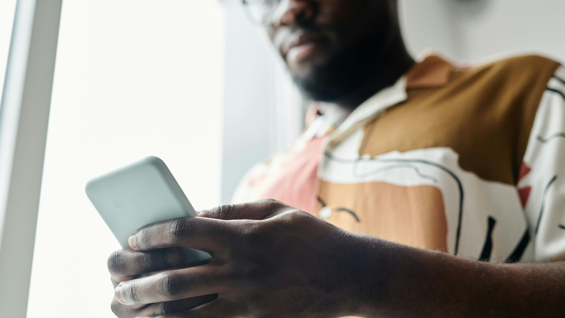 A close-up of a man using a smartphone by a window, featuring a modern patterned shirt.