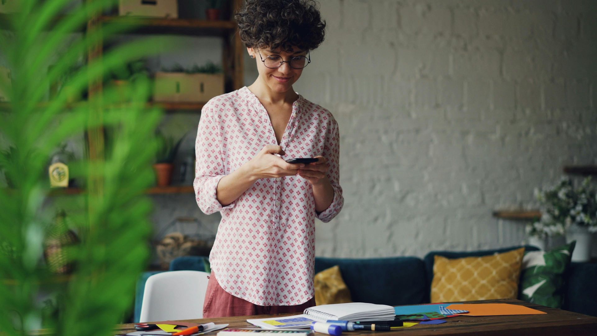 A woman in a casual setting using a smartphone, surrounded by plants and stationery.