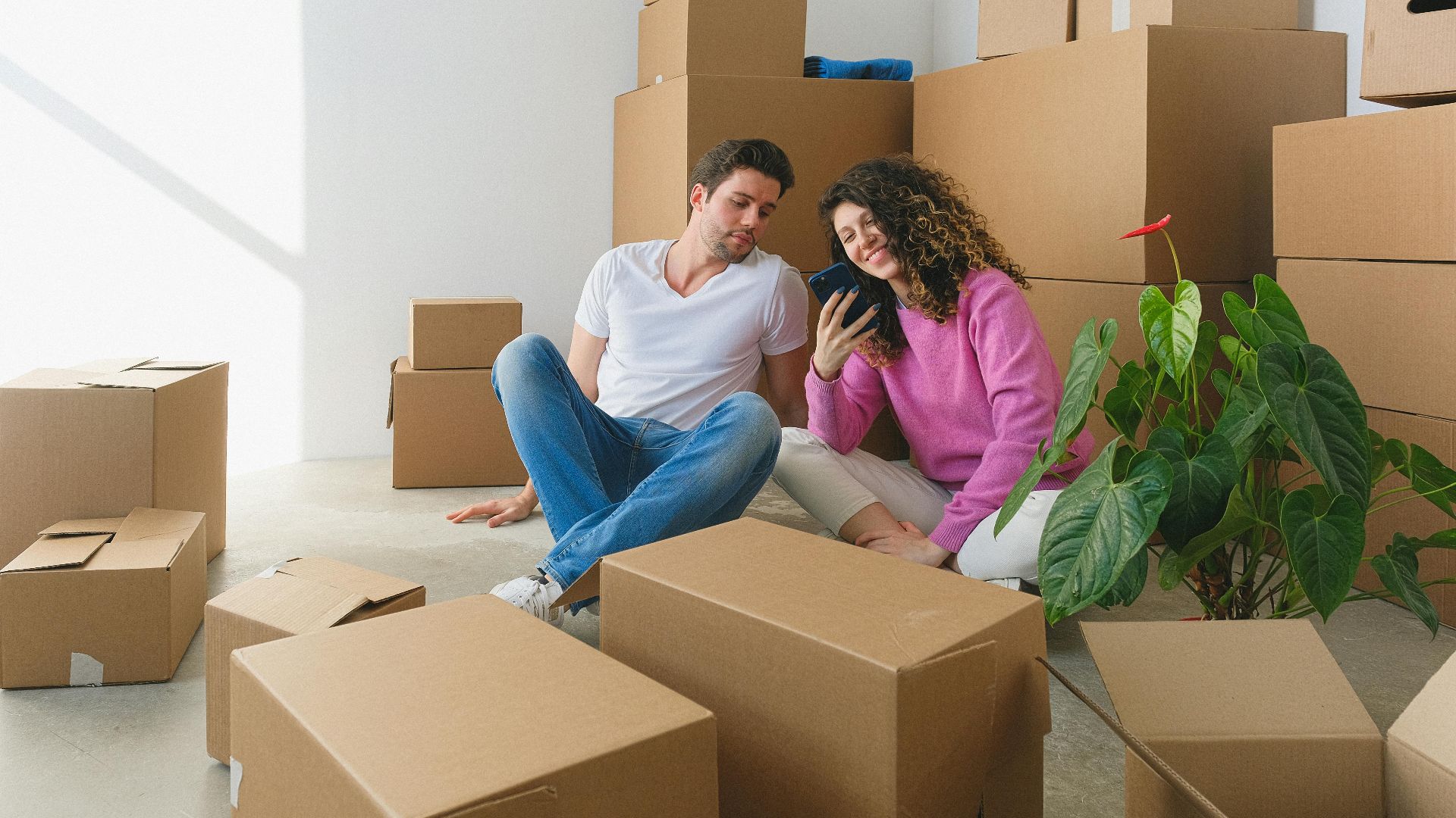 Young couple sitting among cardboard boxes, looking at smartphone, symbolizing moving or relocation.