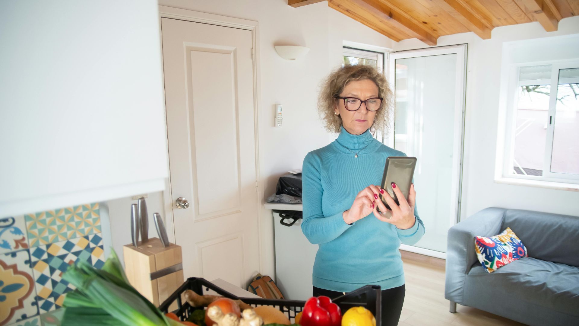 Elderly woman checks her smartphone in a cozy kitchen with fresh vegetables.