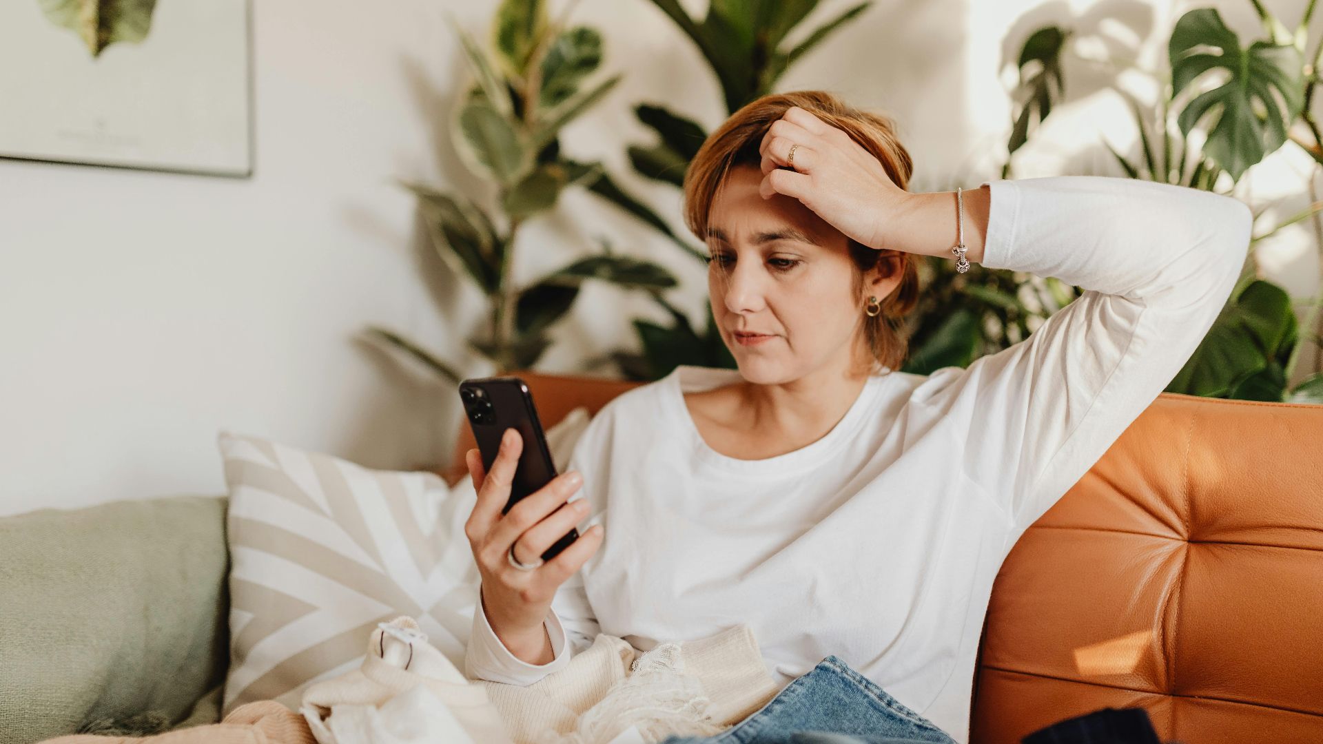 Young woman in white blouse using smartphone while sitting on an orange sofa surrounded by plants.